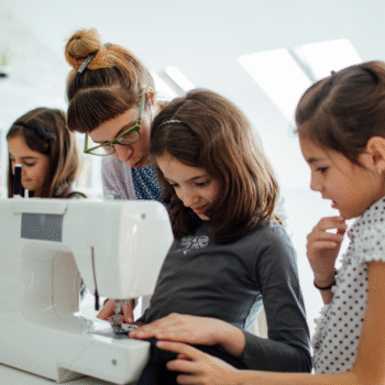 Copy of blog 1 sf (2) A sewing instructor helps a group of young girls learn to sew using a sewing machine in a bright, modern classroom setting.