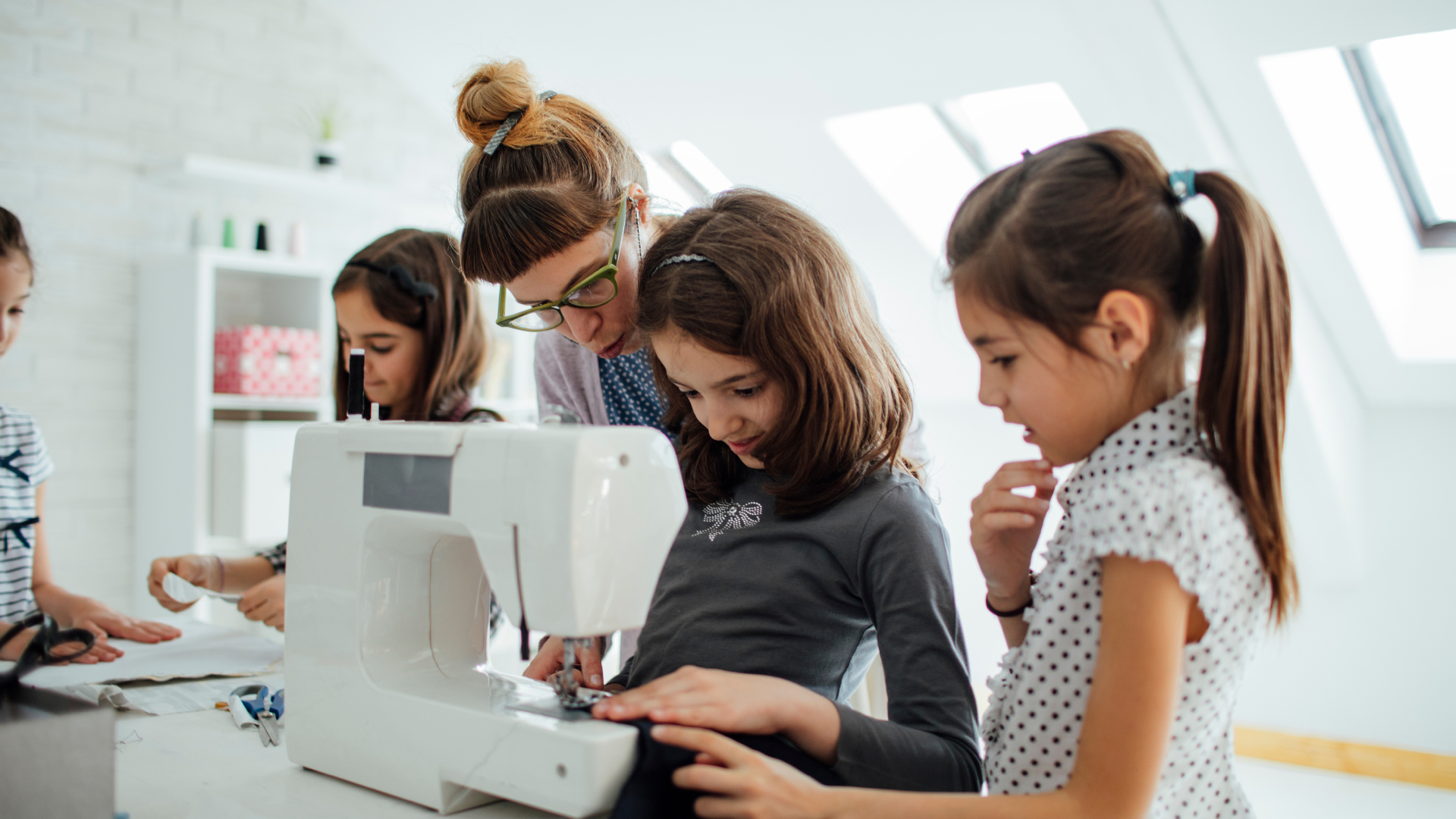 A sewing instructor helps a group of young girls learn to sew using a sewing machine in a bright, modern classroom setting.